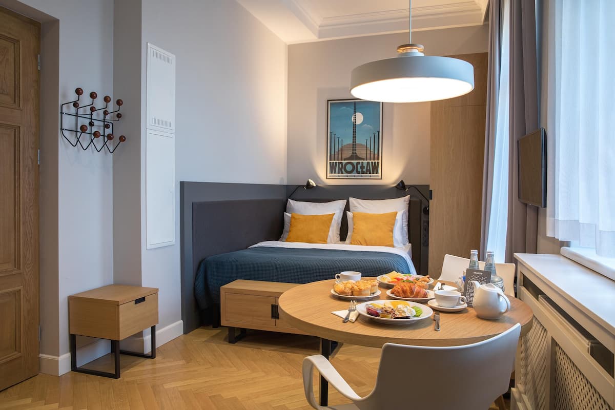 Una habitación de hotel moderna con una cama bien hecha, una mesa de comedor con desayuno, una mesita auxiliar y un gran ventanal con cortinas.