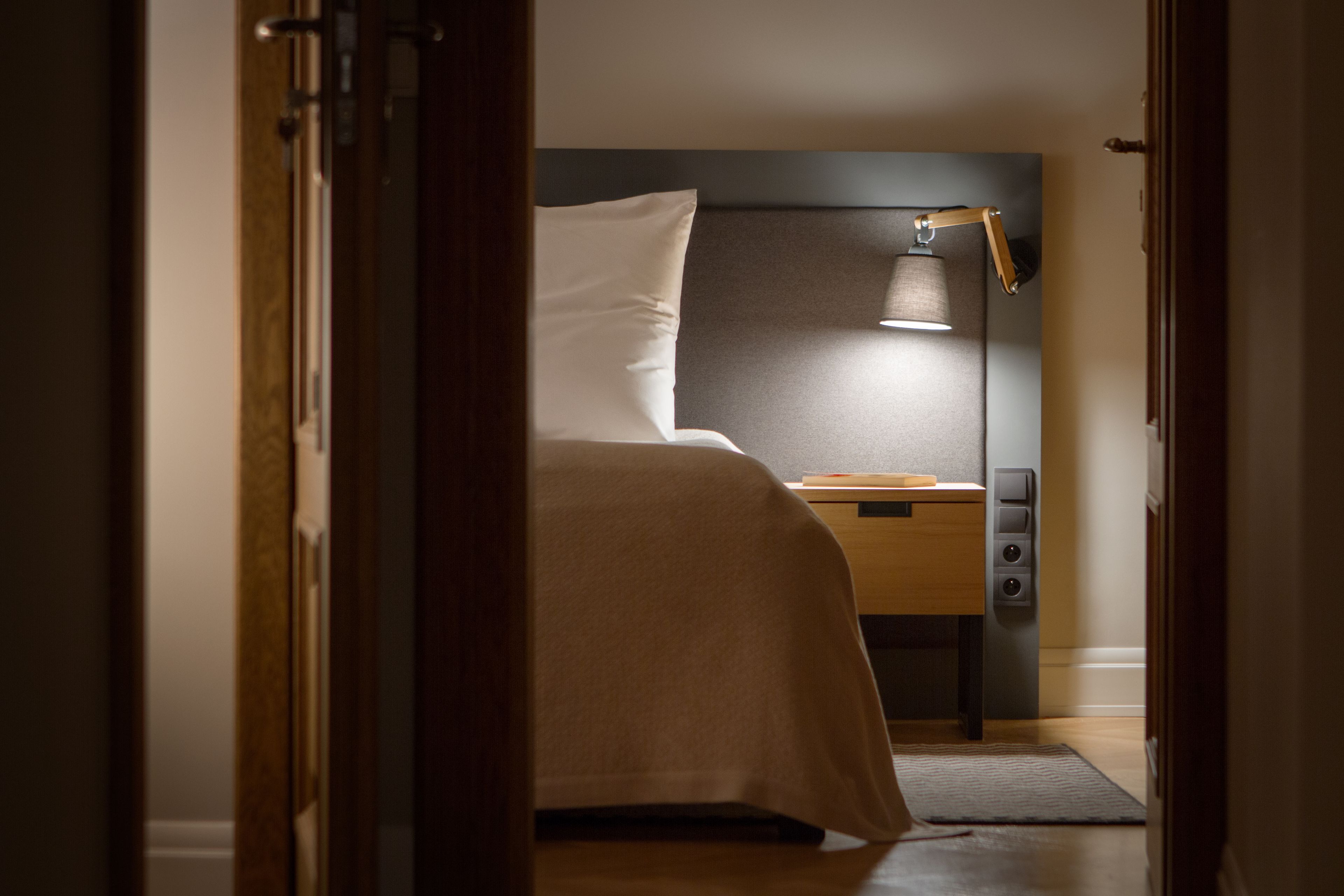 Una camera d'albergo con un letto, un comodino, una lampada e una porta che dà su un'altra stanza.