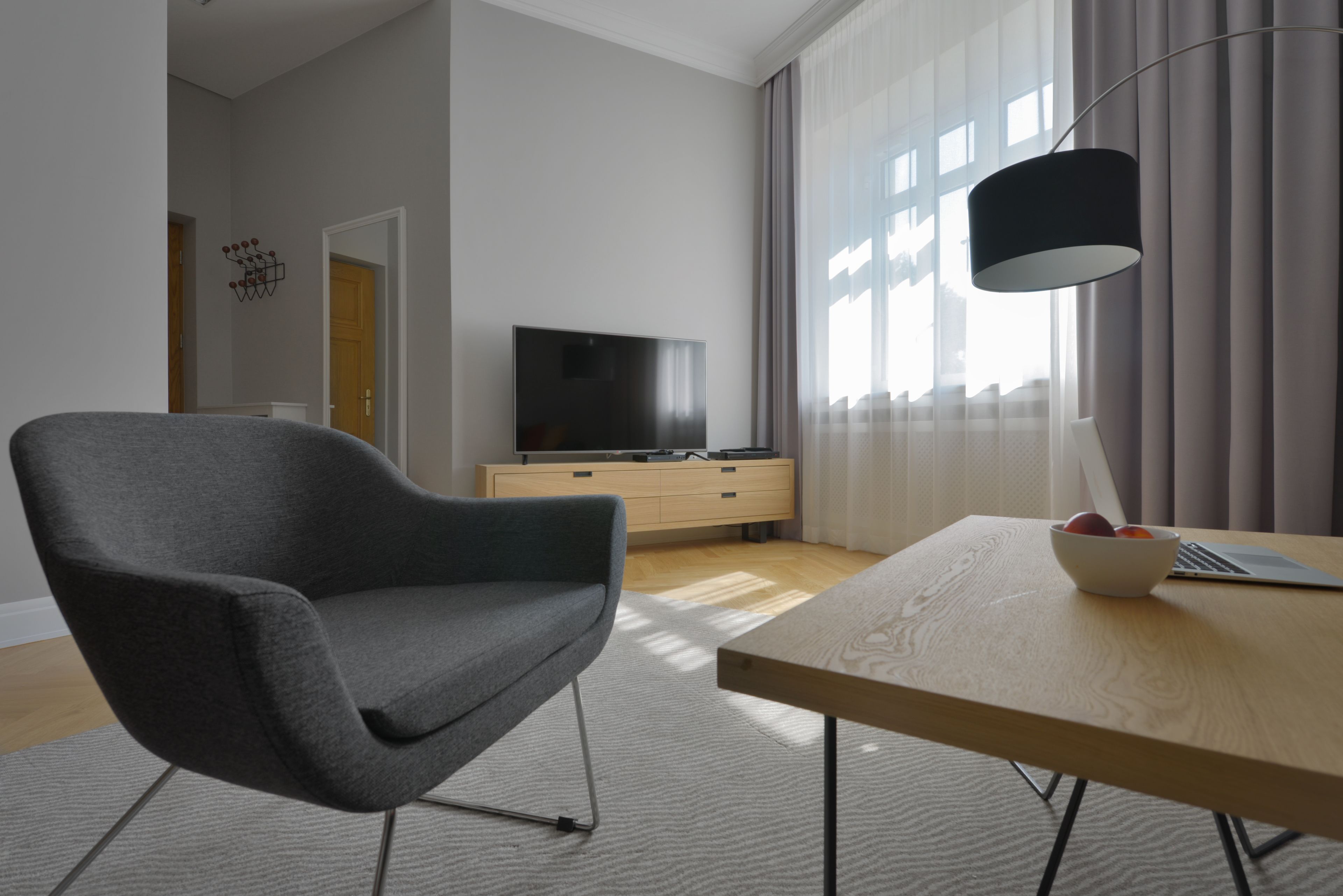 Un salon moderne avec un fauteuil gris, une table basse en bois sur laquelle se trouve un saladier de fruits, un téléviseur posé sur un meuble en bois et un lampadaire noir.