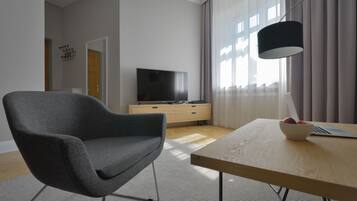 A modern living room with a grey armchair, a wooden coffee table with a bowl of fruit, a TV on a wooden cabinet, and a black floor lamp.