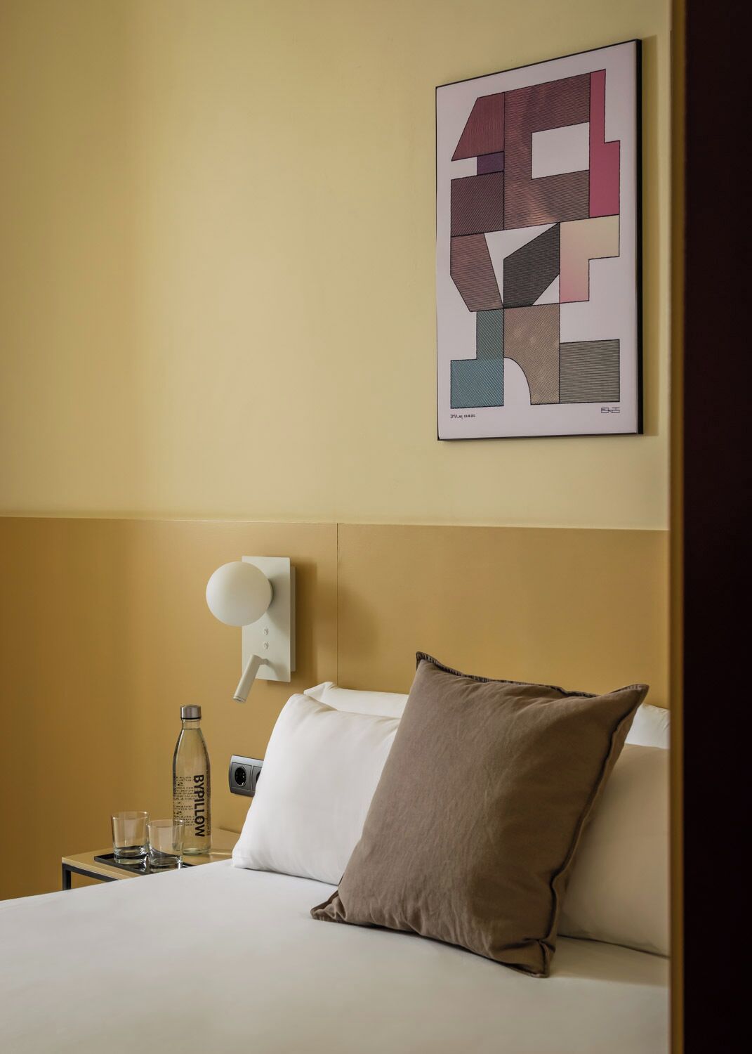 Een hotelkamer met een bed, kussens, een wandlamp, een fles en glazen op een nachtkastje.