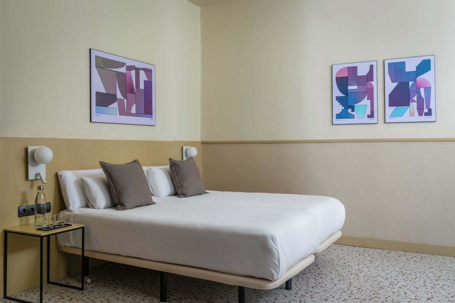 Une chambre d’hôtel avec un lit, deux oreillers, une table de chevet avec une lampe et des œuvres d’art abstrait accrochées aux murs.