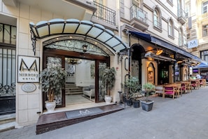 A hotel entrance with a glass canopy, outdoor seating, and a sign for the 'Mirror Hotel.'