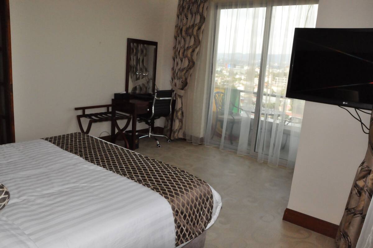 A hotel room with a bed, a desk, a chair, a television, and a view of the city through large windows.