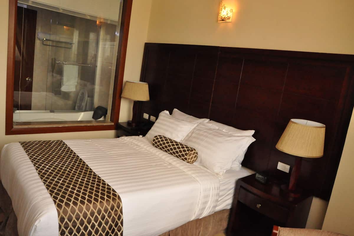 A hotel room with a large bed, a wooden headboard, two bedside lamps, a window with a view of a shower area, and a wall-mounted light fixture.