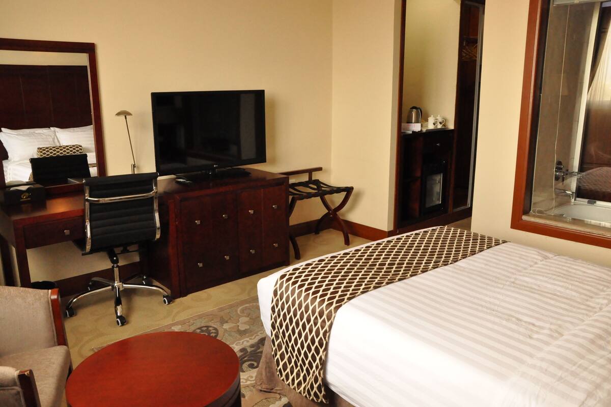 A hotel room with a bed, a desk with a TV, a chair, and a small table.