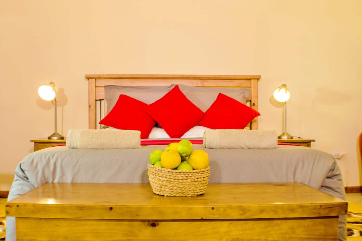 Un letto rifatto con cuscini rossi, un comodino con un cesto di frutta e due abat-jour.