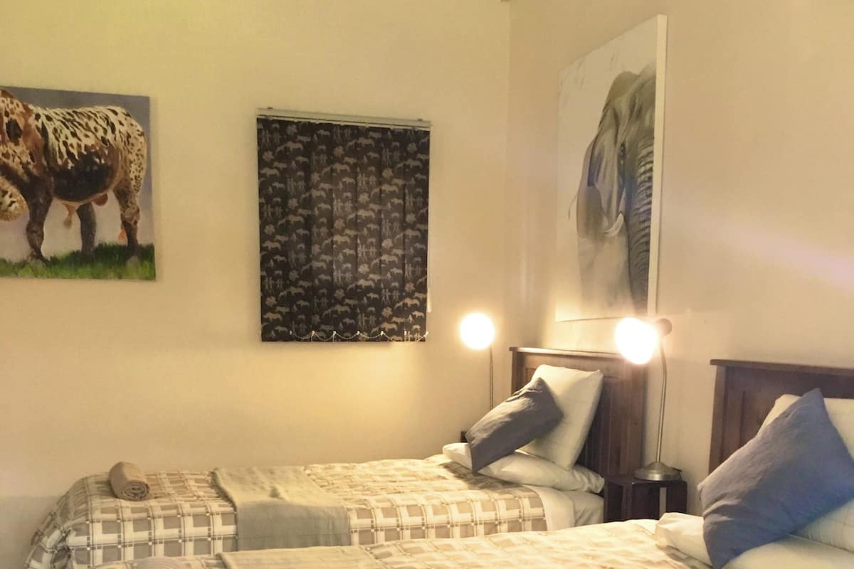 Un letto matrimoniale con copriletto e cuscini a quadri, un comodino con una lampada, e un muro con un dipinto che raffigura una mucca e una foresta.