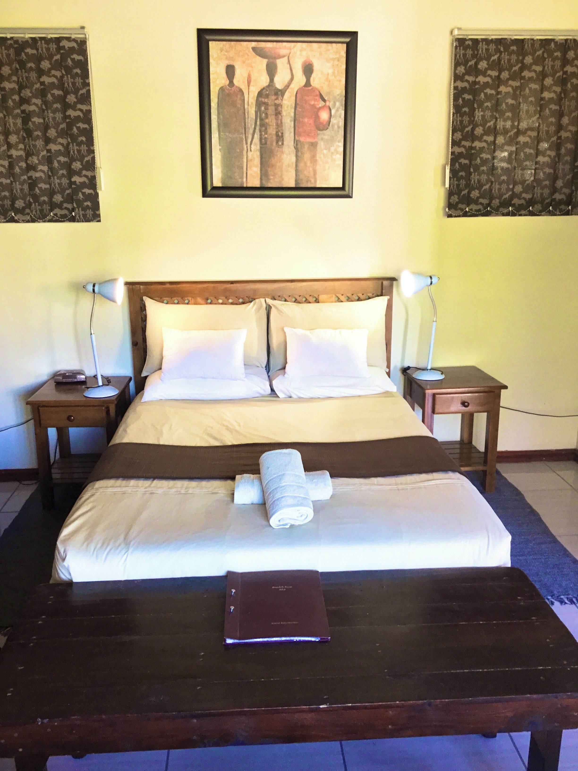 A neatly made bed with white linens, two bedside tables with lamps, a painting on the wall, and a wooden table with a book in the foreground.