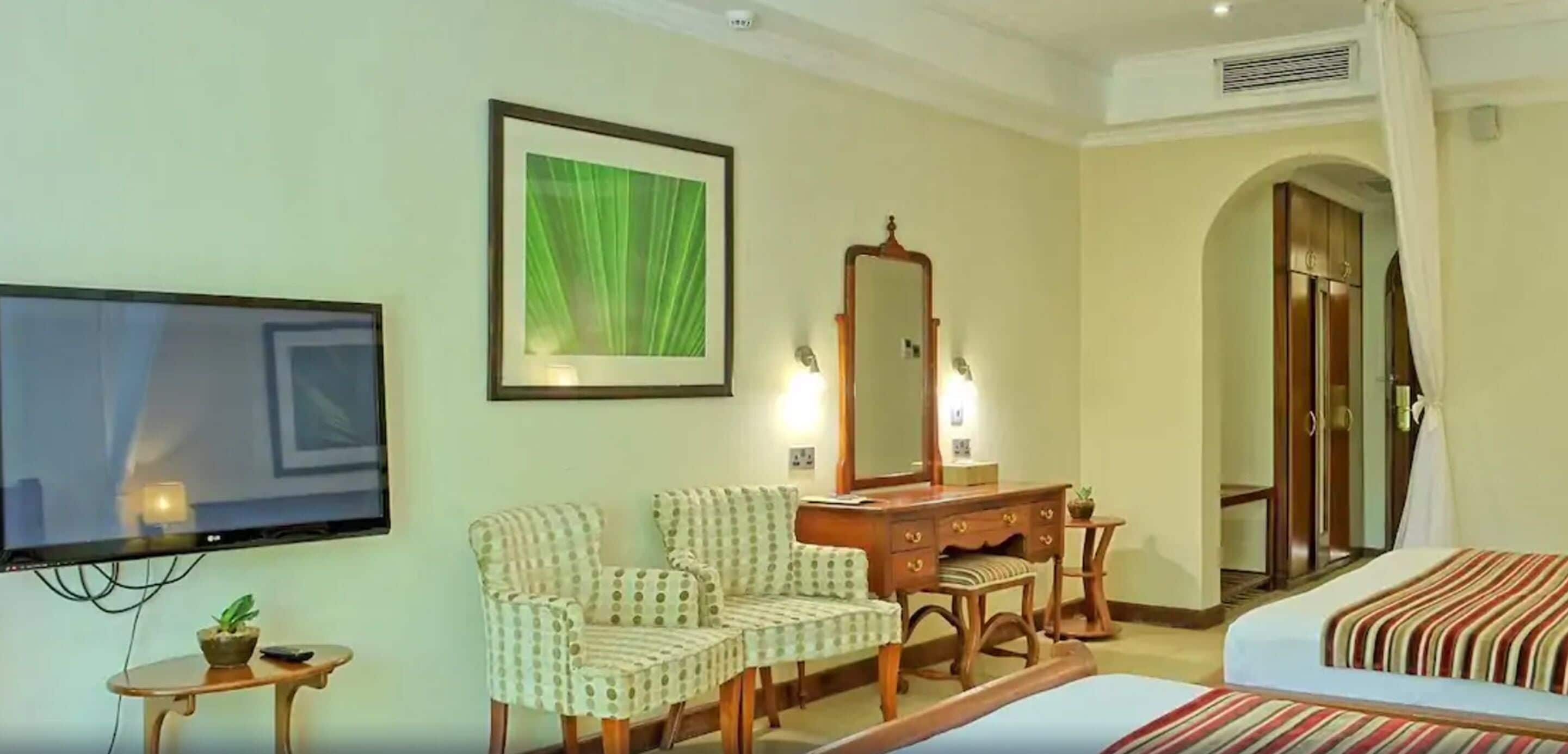 Habitación de hotel con televisión, dos sillones estampados, cómoda de madera, espejo y cama con ropa de cama a rayas.