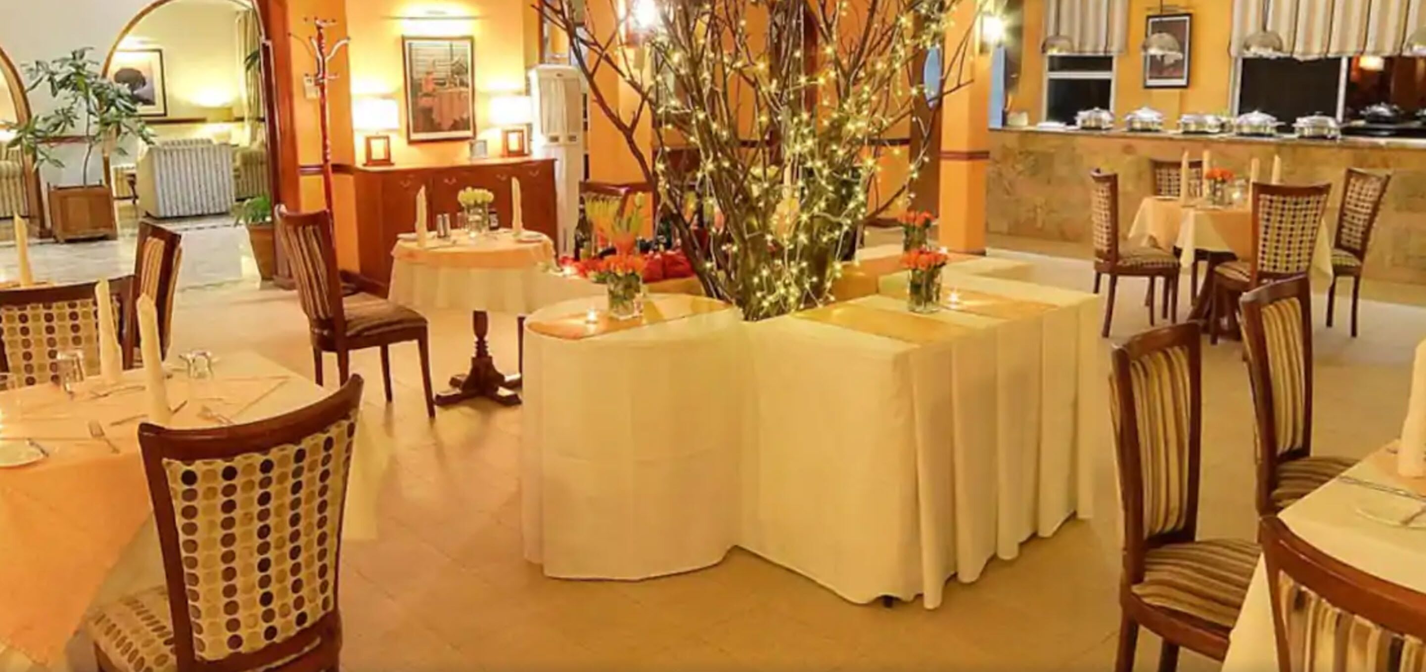 Zona de menjador amb taules rodones, cadires i un arbre decoratiu al centre.