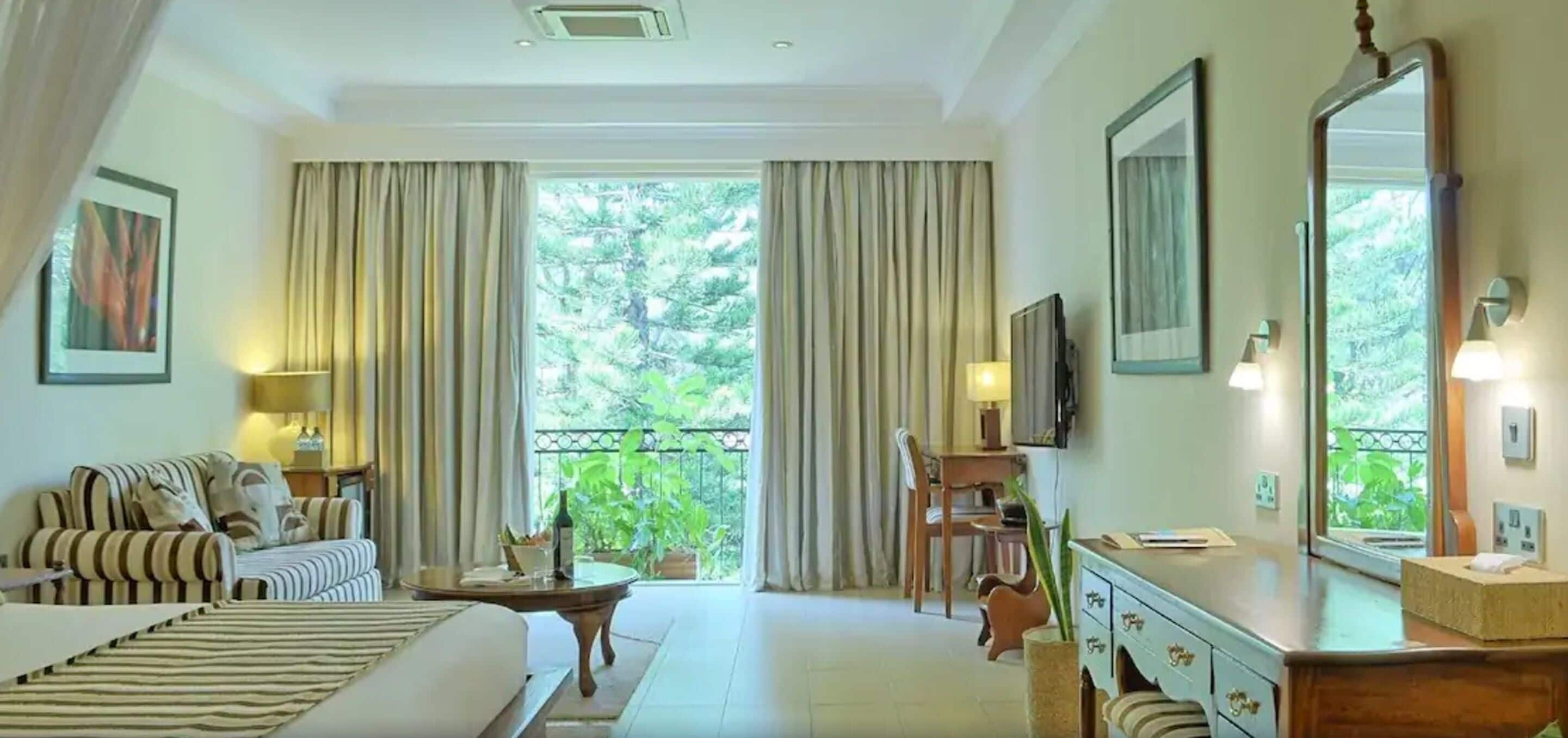 Habitación de hotel con cama, sofá, mesa de comedor, escritorio y un amplio ventanal con cortinas.