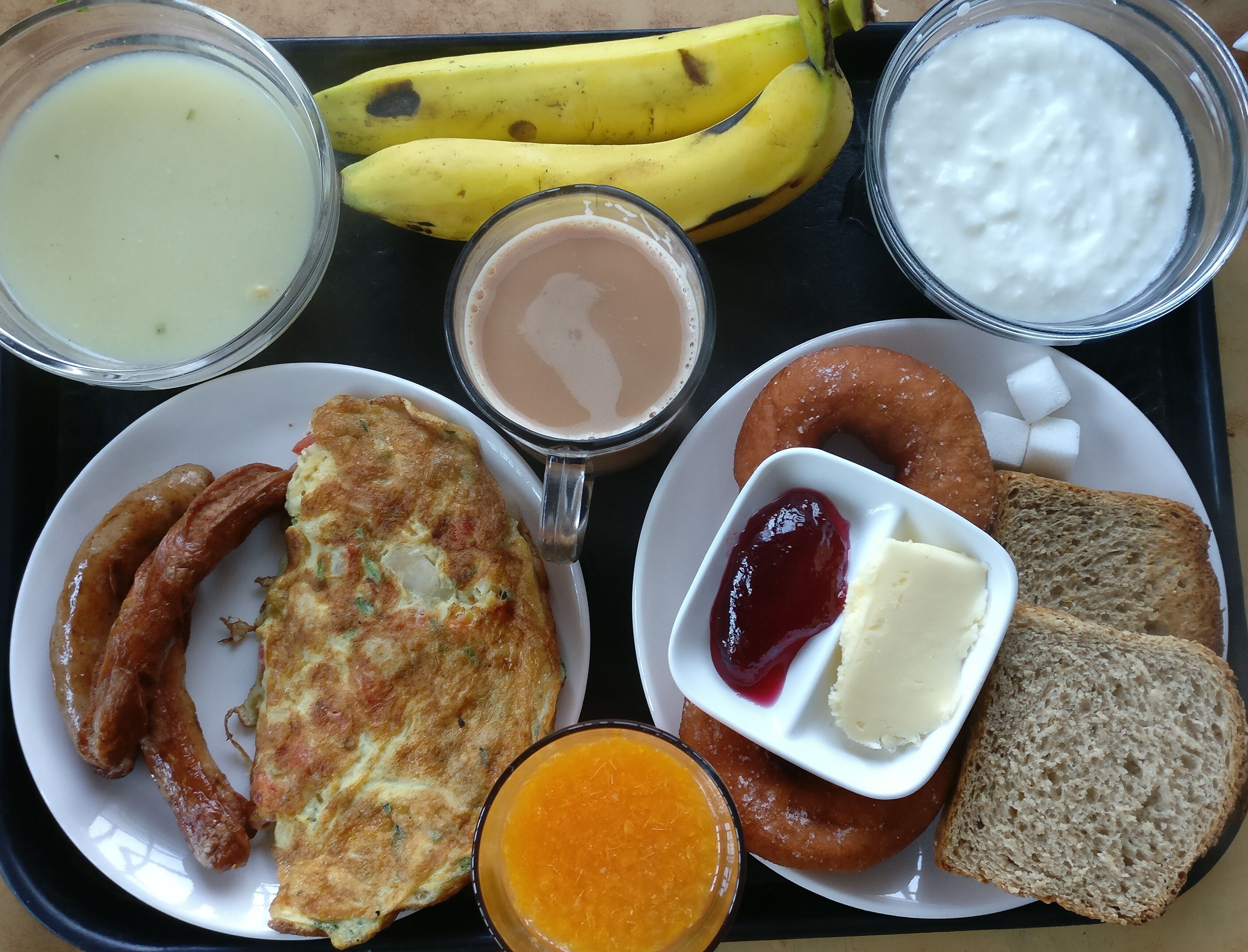 Una bandeja con alimentos para desayuno, incluyendo salchichas, una tortilla, tostadas, mermelada, mantequilla y un bol de jugo de naranja.