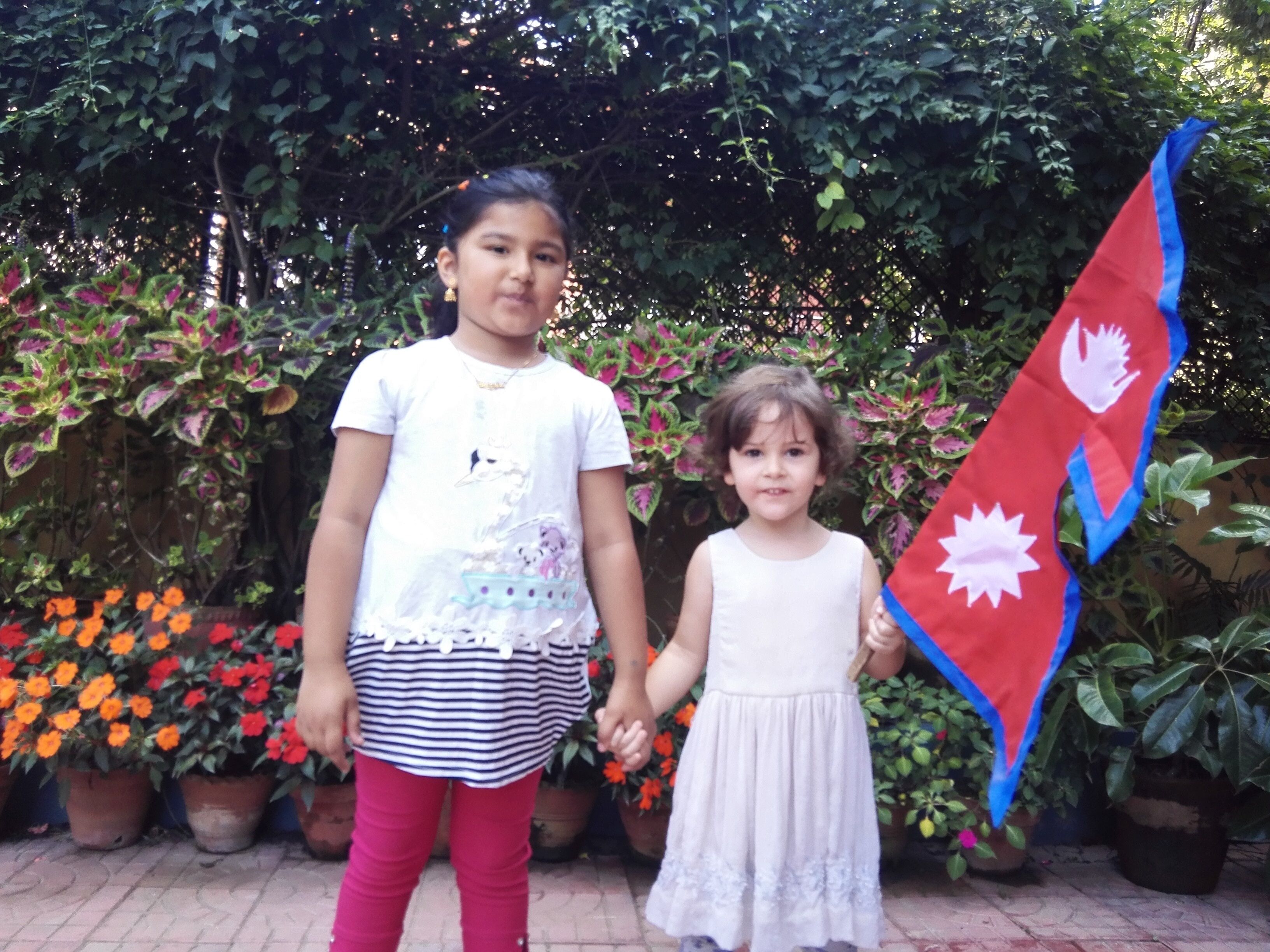 Two children holding hands in a garden with potted plants and flowers.