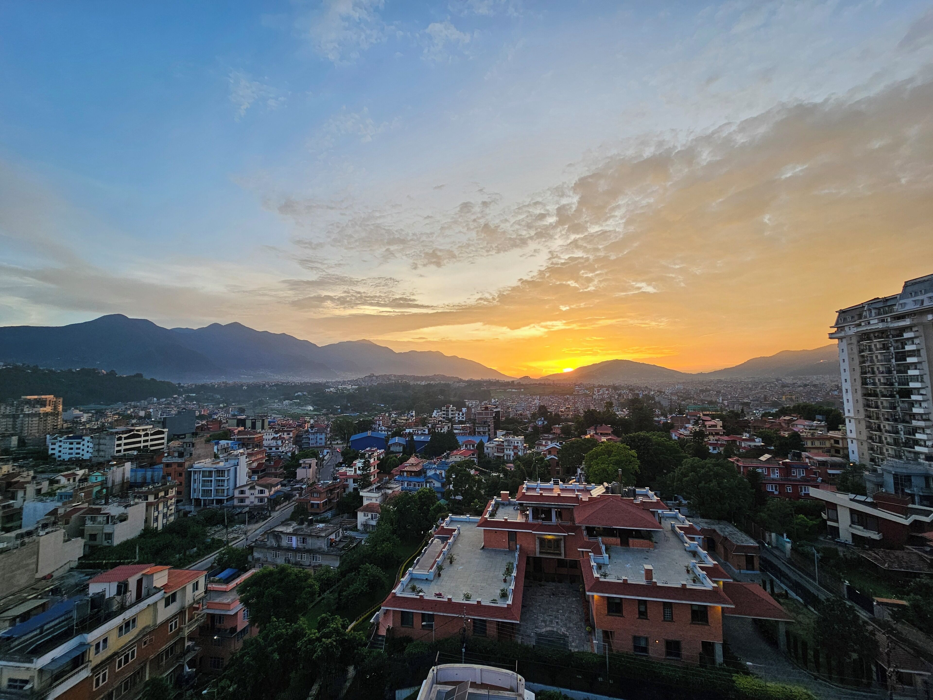 Une vue urbaine avec un coucher de soleil sur des montagnes, mettant en scène des immeubles résidentiels dotés de toits plats et présentant un mélange de styles architecturaux.