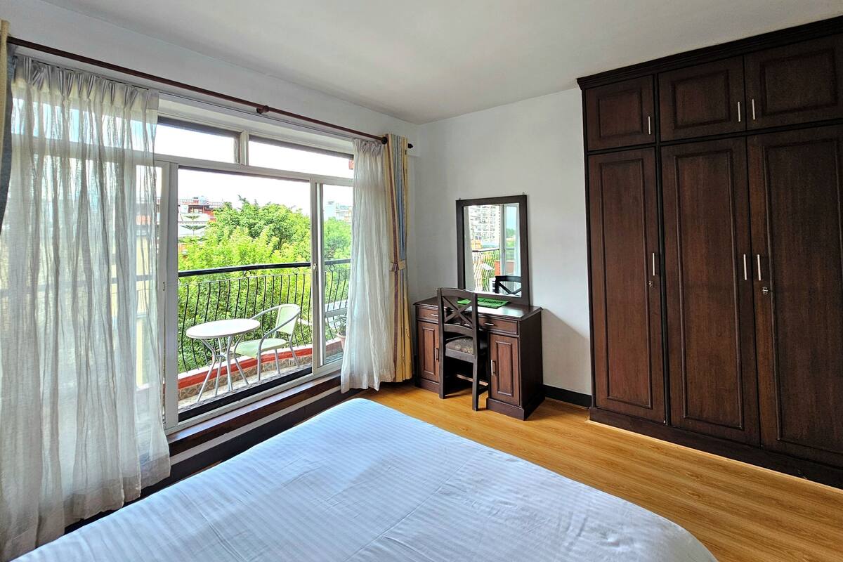 Une chambre dotée d’un grand lit, d’un bureau avec une chaise, d’une armoire et d’un balcon offrant une vue sur la verdure.