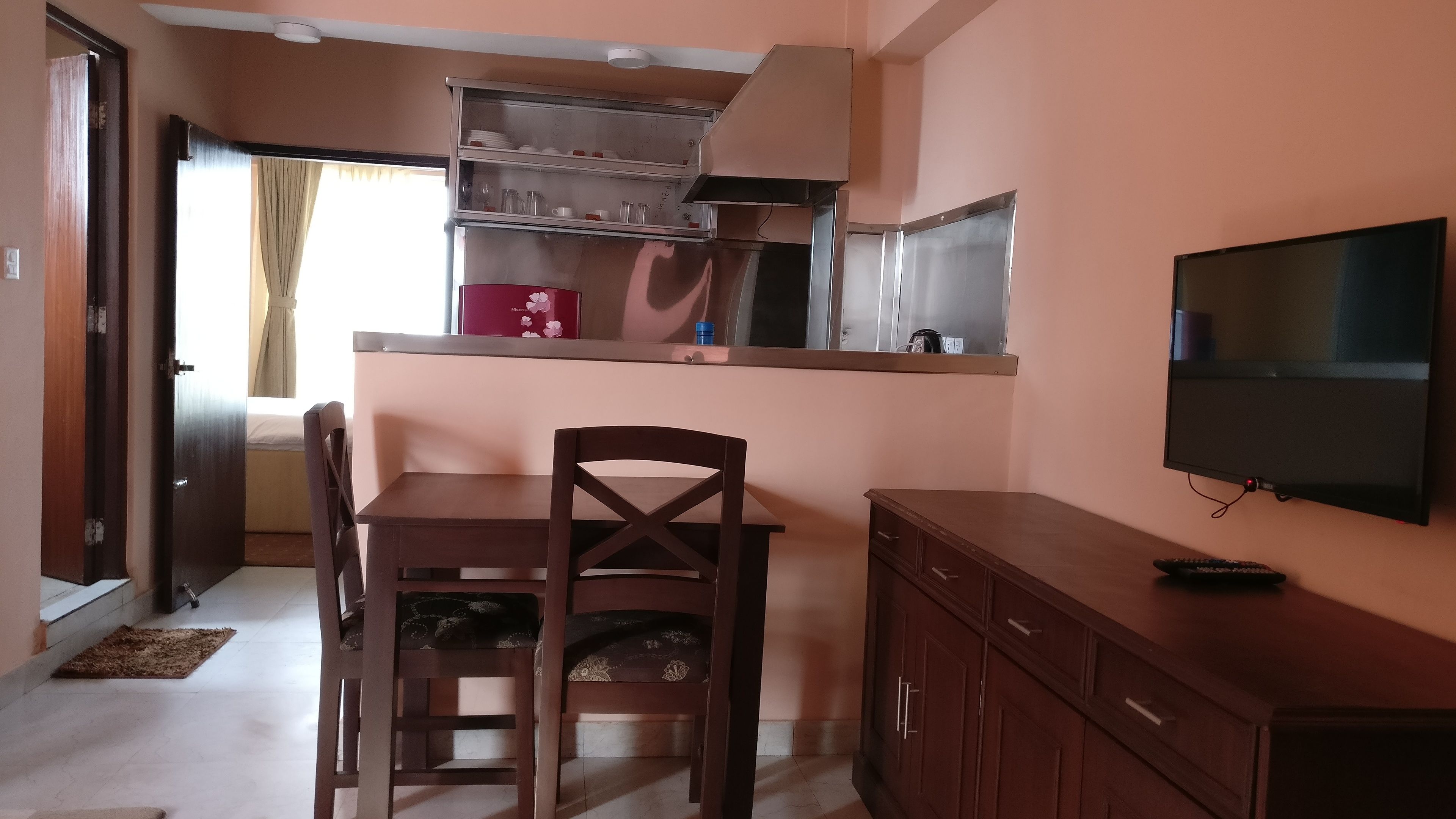 A compact kitchen with a dining table and chairs, a TV mounted on the wall, and a window with curtains.