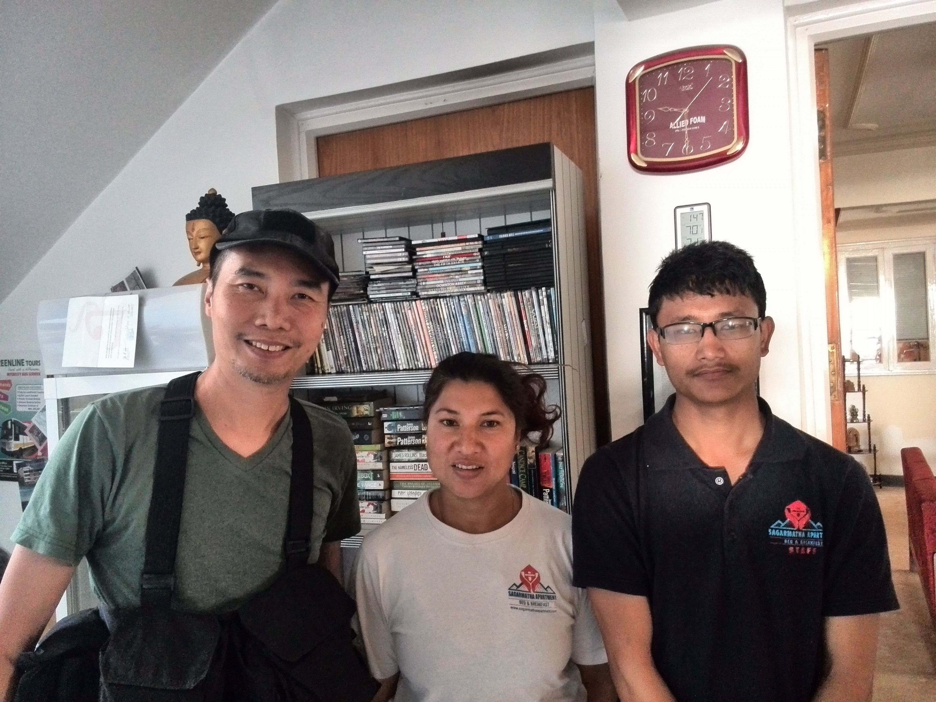 Three people standing in a room with a bookshelf filled with DVDs and a clock on the wall.