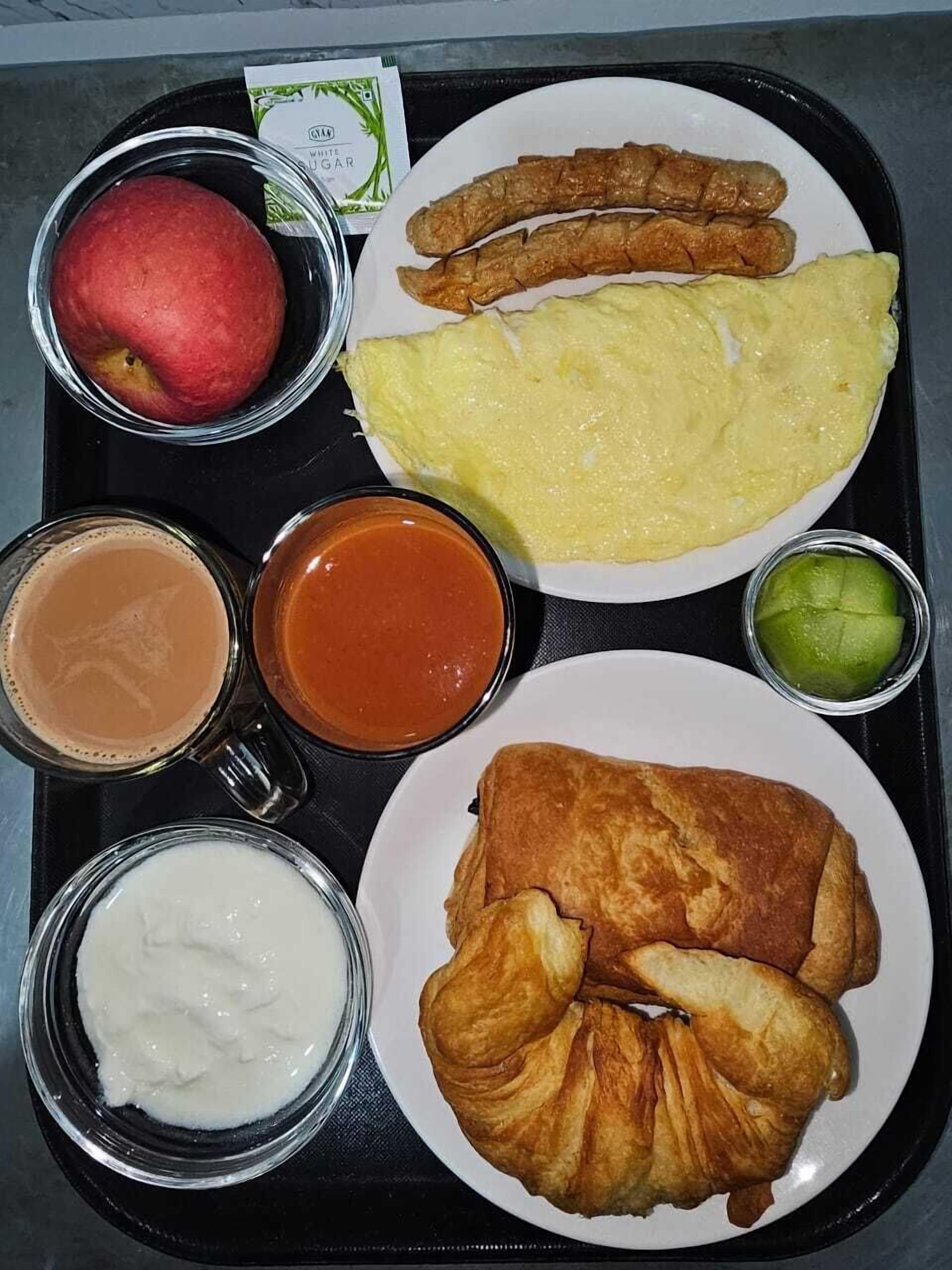 A tray with a plate of food including an omelette, sausage, croissant, and apple, along with three cups of beverages.