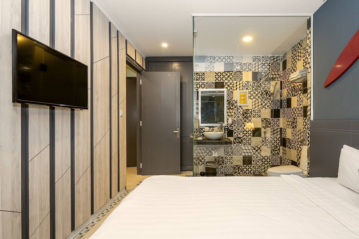 Une chambre d’hôtel moderne dotée d’un grand lit, d’un téléviseur fixé au mur, d’une salle de bain aux carreaux décoratifs et d’une douche à parois de verre.