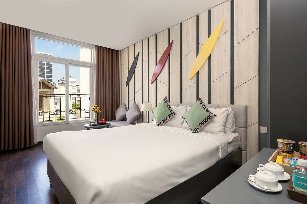Une chambre d’hôtel avec un grand lit, une petite table sur laquelle se trouvent un vase et un panier de fruits, et une vue sur les bâtiments par la fenêtre.