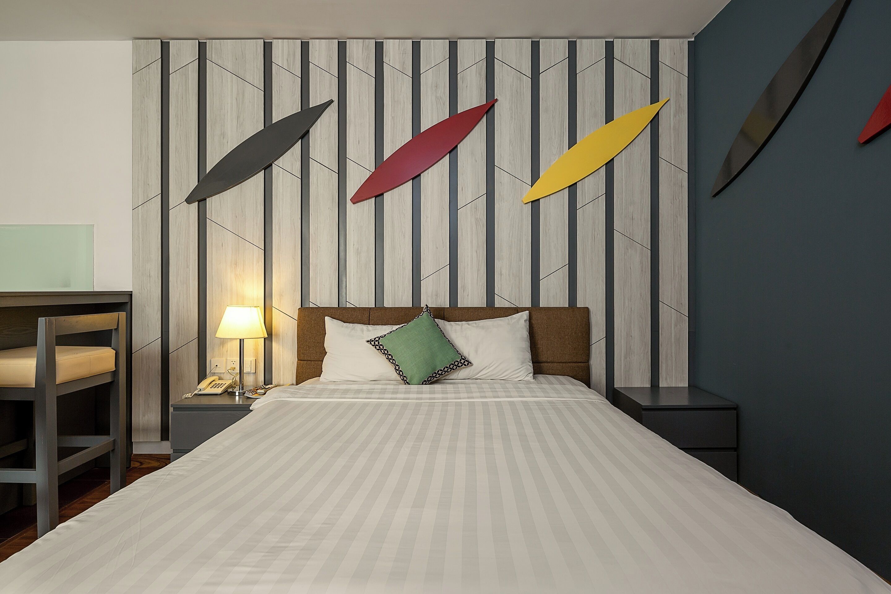 A neatly made bed with a striped bedspread, a bedside table with a lamp, and a wall with geometric patterns.