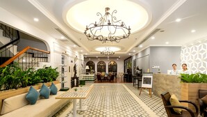 Un hall d’entrée spacieux doté d’un sol à motifs géométriques, d’un lustre central, d’un comptoir de réception et d’un espace détente avec un canapé et des fauteuils.