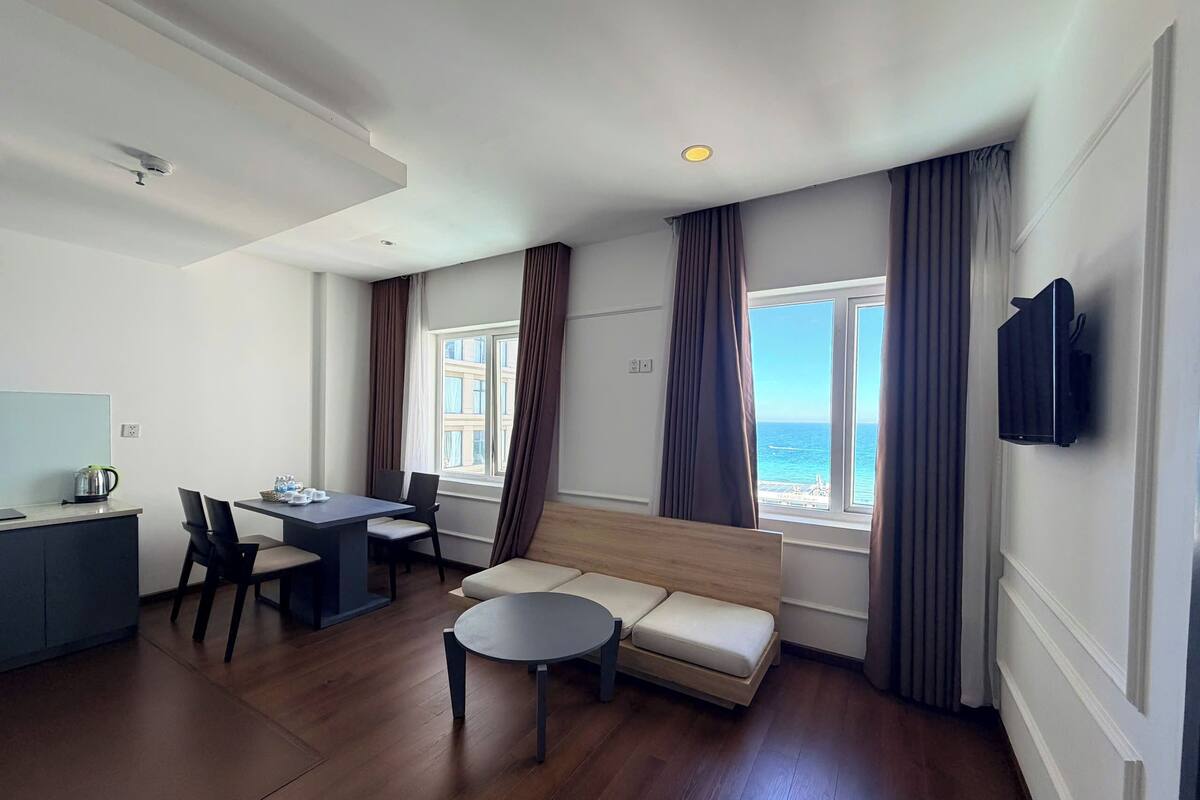 Une chambre d’hôtel moderne avec un plancher en bois, un canapé, une table à manger avec des chaises, un coin cuisine et une grande fenêtre donnant sur la mer.