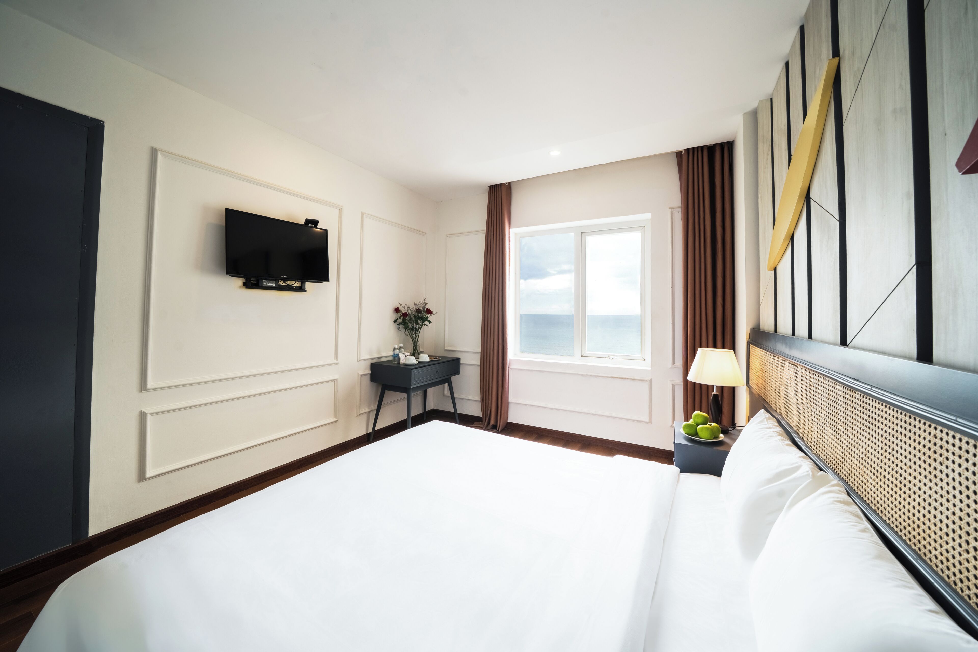 Une chambre d’hôtel avec un grand lit, un téléviseur à écran plat fixé au mur, une petite table surmontée d’un vase de fleurs, une fenêtre avec des rideaux et une table de chevet avec une lampe et des pommes.