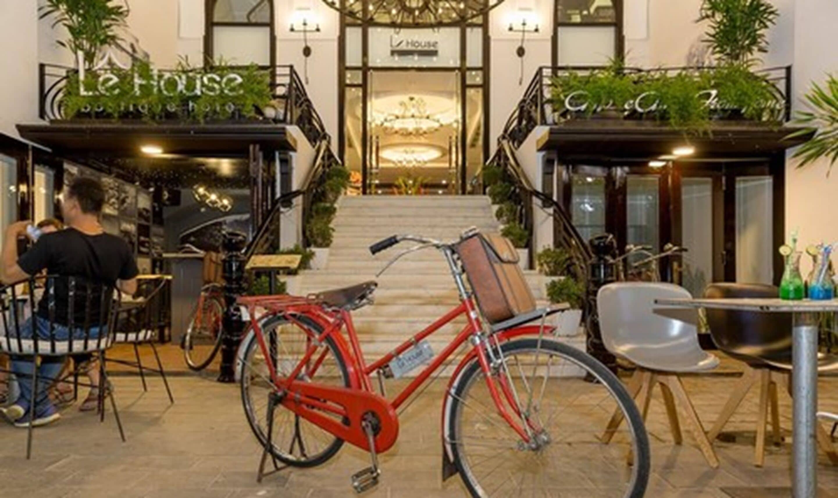 A hotel entrance with a staircase leading to the lobby, a red bicycle parked in the foreground, and a person sitting at a table.