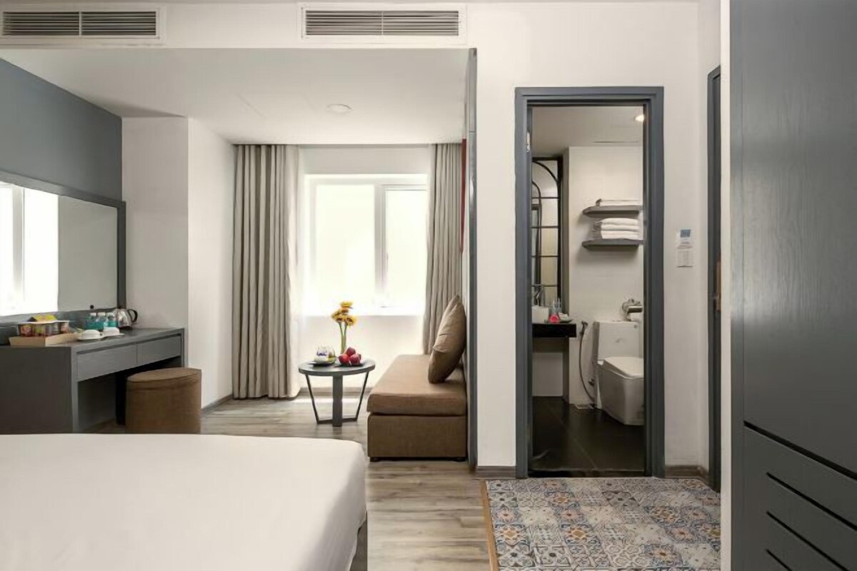 Ein Hotelzimmer mit einem Bett, einem Schreibtisch, einem Stuhl, einem kleinen Tisch mit einer Vase und einem durch eine offene Tür einsehbaren Badezimmer.