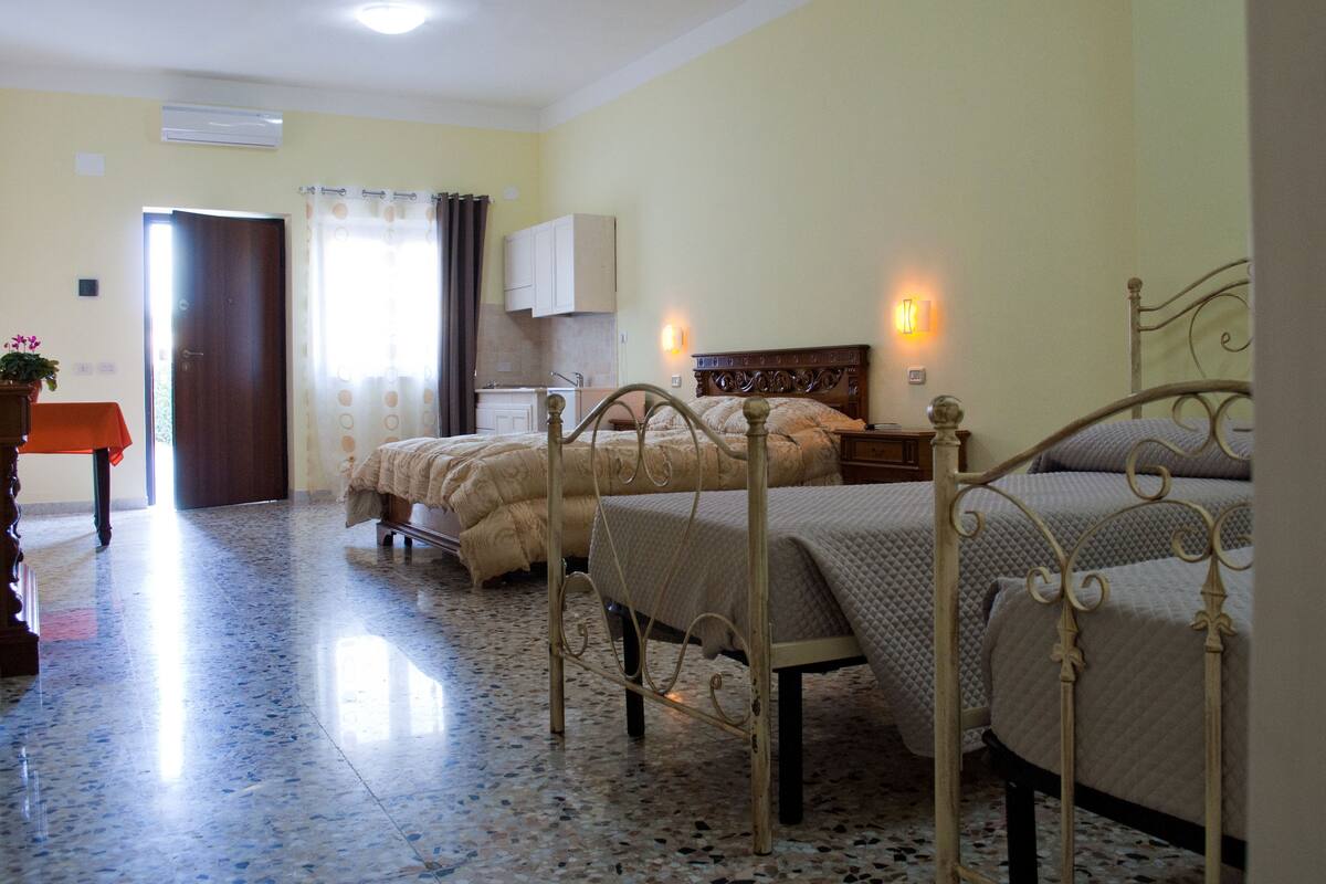 Una camera d'albergo con tre letti, un tavolino con una pianta, angolo cottura e bagno con tenda doccia.