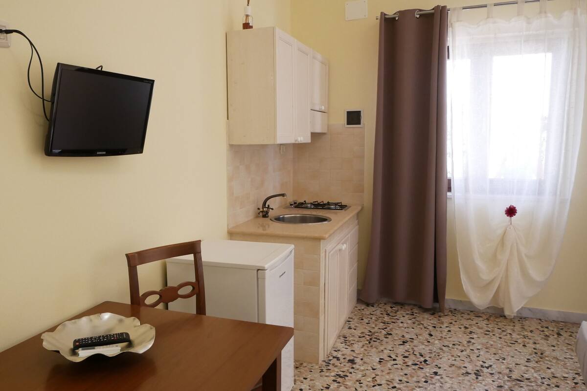 Una cucina compatta con lavello, piano cottura e pensili, un tavolo da pranzo con telecomando e un televisore montato a parete.