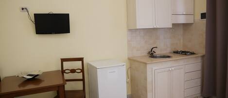 A compact kitchen with white cabinets, a small refrigerator, a sink with a faucet, and a television mounted on the wall.