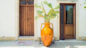 Un jarrón naranja grande con una planta de cactus sobre un piso de baldosas, junto a una puerta de madera y una ventana con contraventanas de madera.