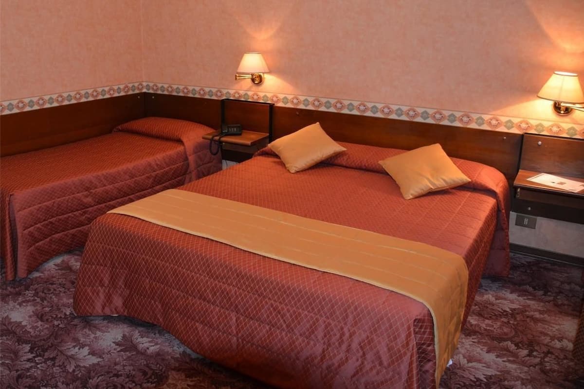 A hotel room with two beds, each with a red bedspread and a beige blanket. There are two bedside tables with lamps, and the room has a carpeted floor.