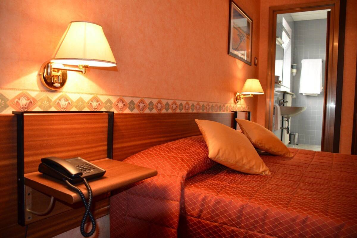 A hotel room with a wooden headboard, a bedside telephone, two pillows, and a wall-mounted lamp.