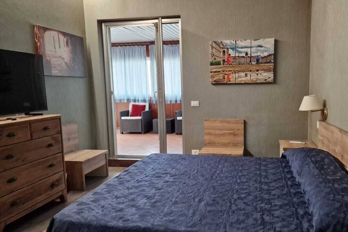 A hotel room with a bed, a wooden dresser, a television, a painting, and a view of the outside.
