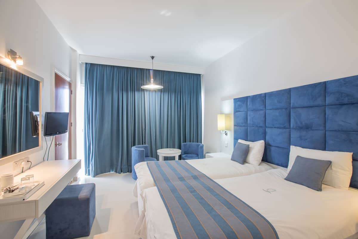 Habitación de hotel con dos camas, un escritorio, una silla, un televisor y un ventanal con cortinas.
