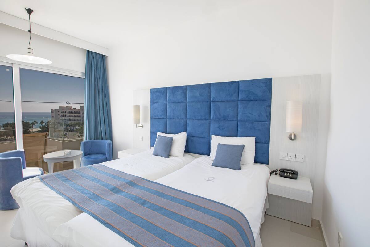 Habitación de hotel con dos camas, vista a la ciudad, balcón y un teléfono en la mesita de noche.
