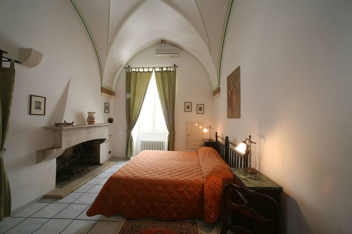A bedroom with a fireplace, a bed with an orange quilt, a desk with a lamp, and a window with curtains.