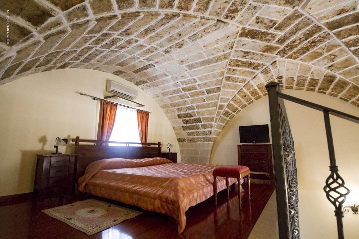 A bedroom with a vaulted stone ceiling, a bed with a red bedspread, a wooden nightstand, a desk with a lamp, and a television on a dresser.