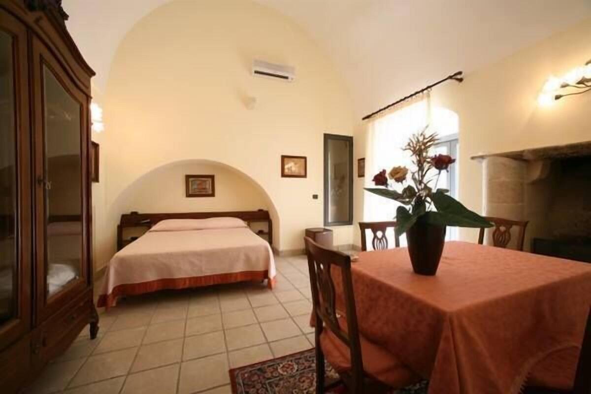 A bedroom with a bed, a wooden wardrobe, a dining table with chairs, and a fireplace.