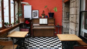Une pièce avec un sol à carreaux, un piano, des tables et des chaises en bois. On y trouve également des plantes en pot et un mur en briques rouges.