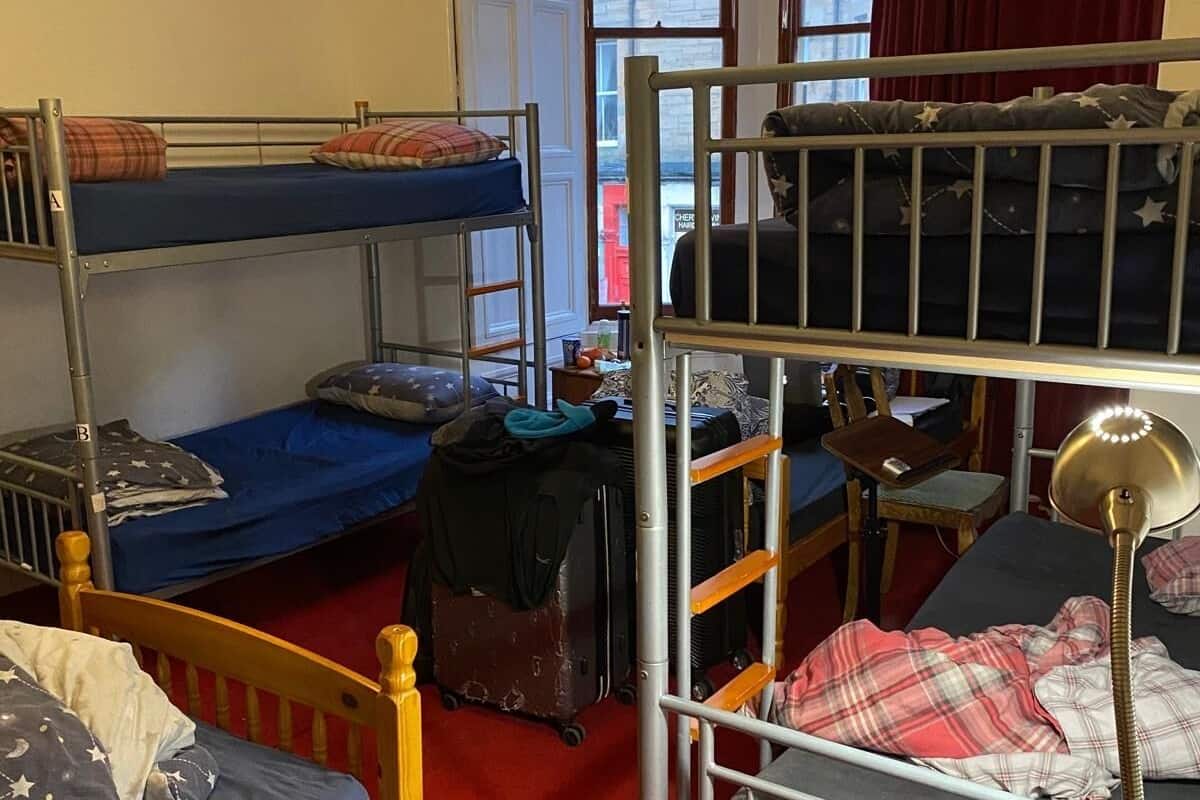 A dormitory room with bunk beds, a suitcase, a lamp, and a window with a view of buildings.