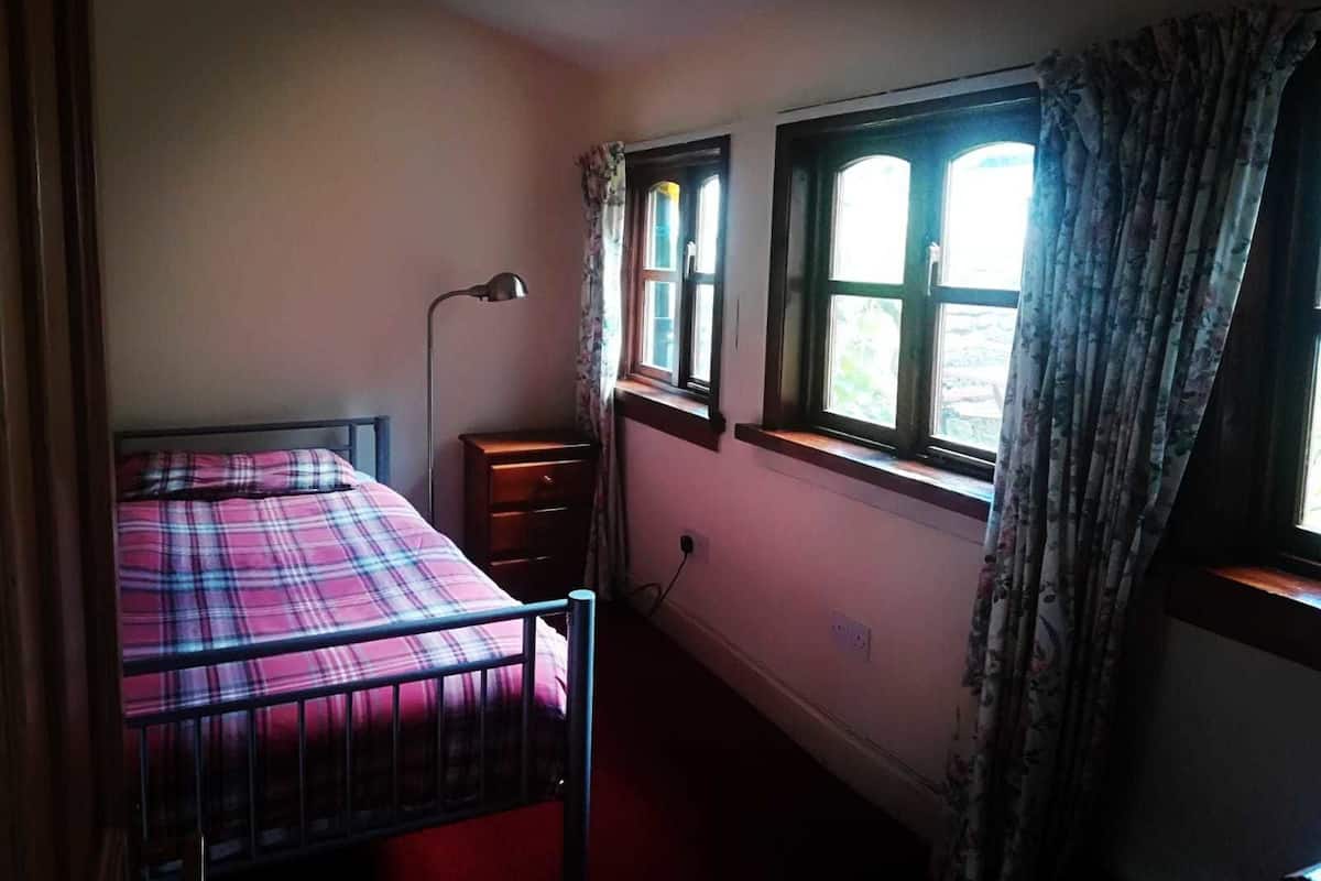 A bedroom with a single bed, a bedside lamp, a nightstand, and a window with curtains.