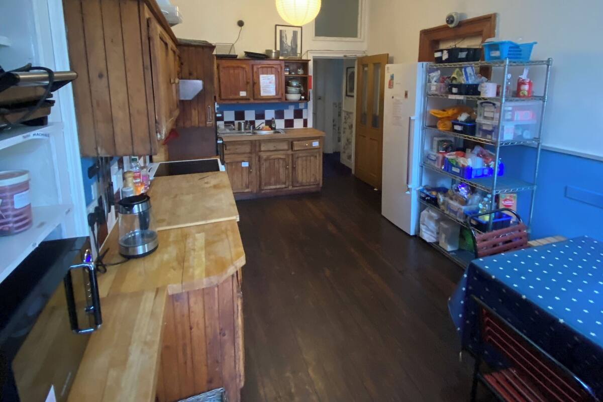 A compact kitchen with wooden cabinets, a wooden countertop, a refrigerator, and a shelving unit with various items.