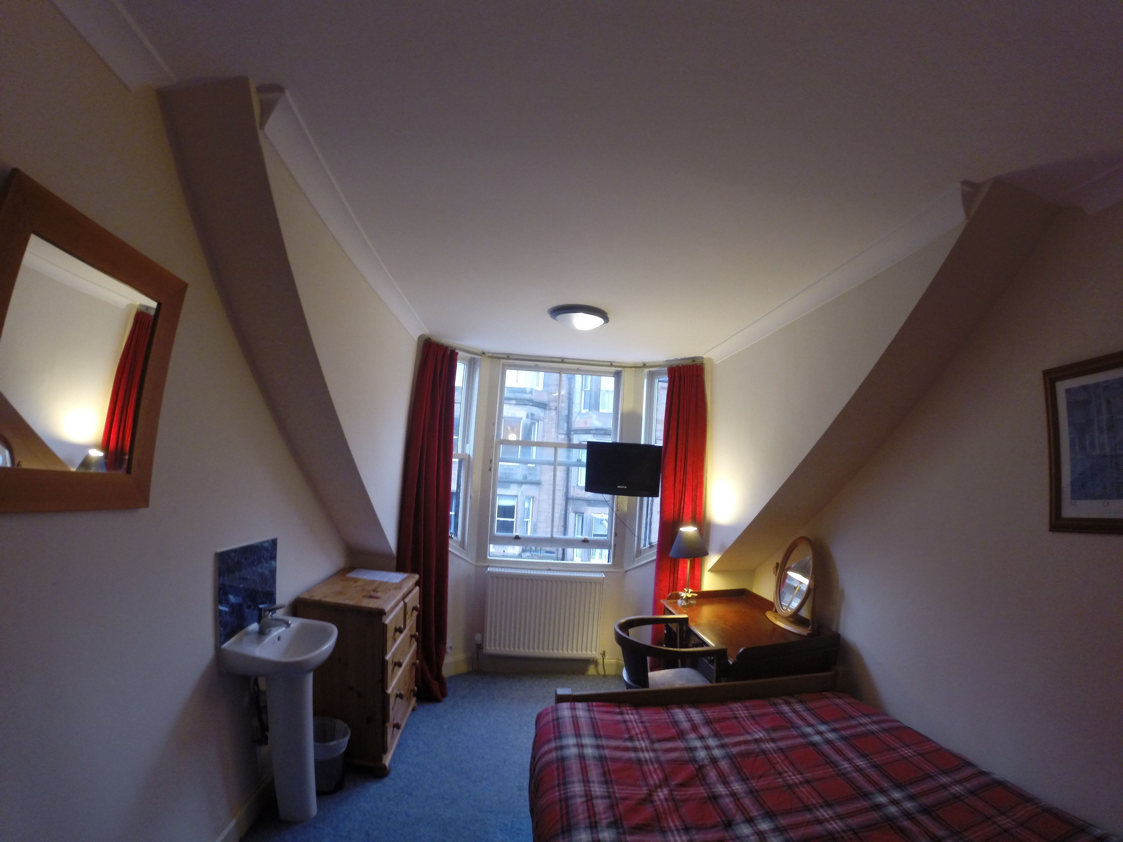 A bedroom with a bed, a desk, a chair, a mirror, a window with curtains, a radiator, and a television.