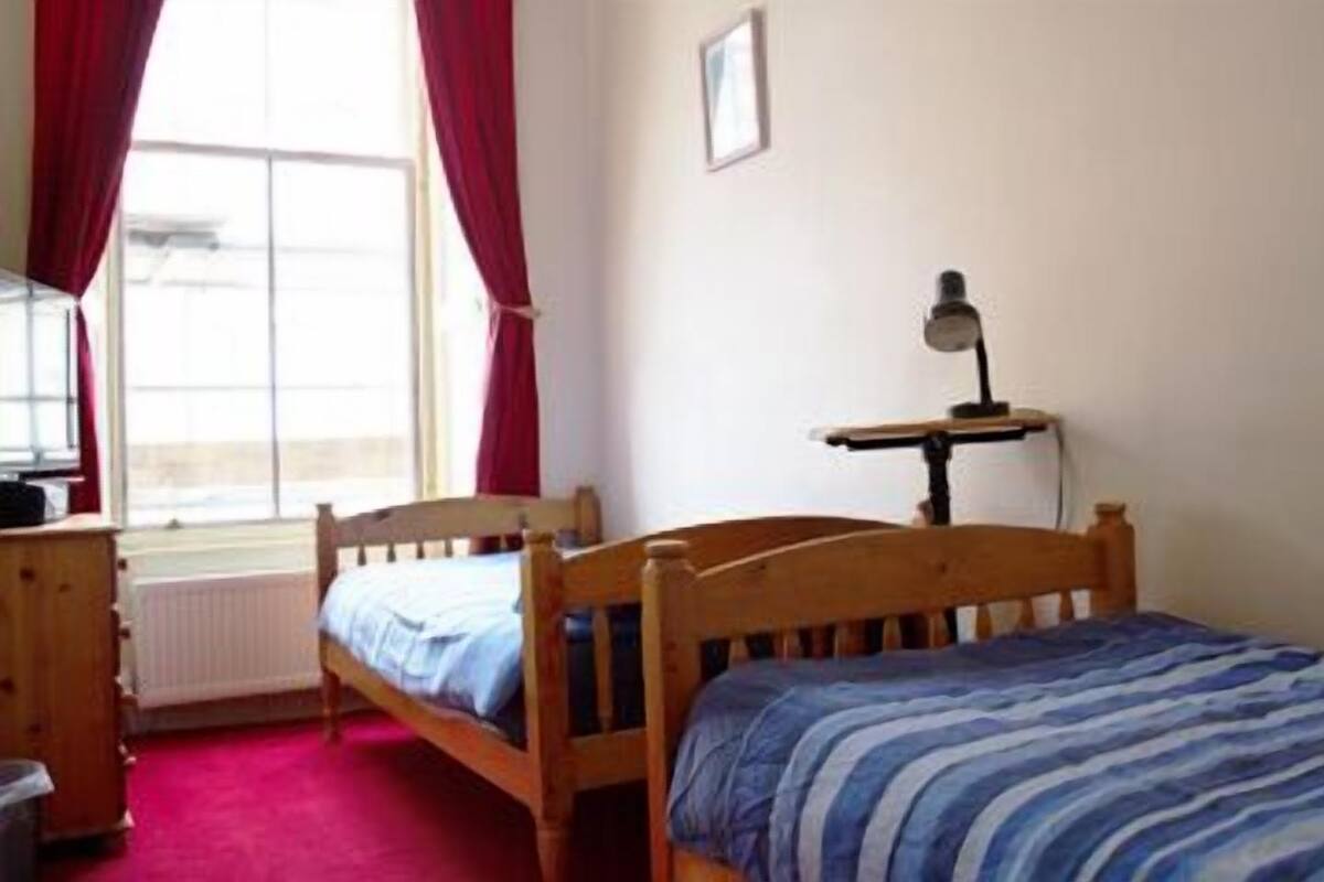 A room with two wooden beds, a desk, a radiator, and a window with red curtains.