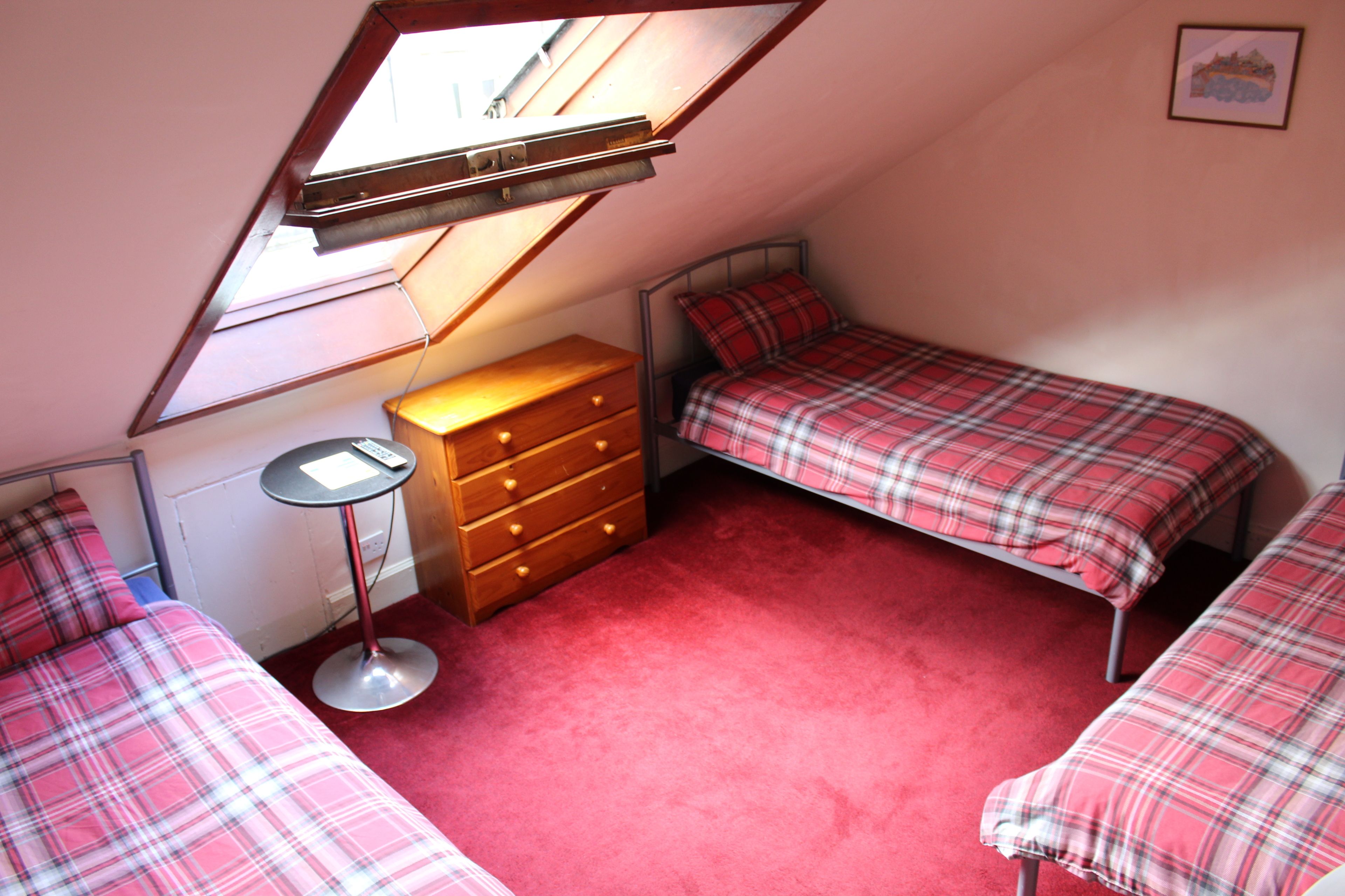 Une petite chambre mansardée de forme triangulaire, avec deux lits, une commode et une petite table.